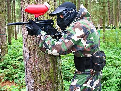 A person wearing camouflage overalls and a mask aiming a paintball marker from behind a tree