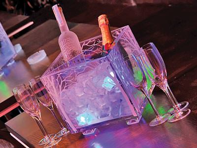 A clear ice bucket containing a bottles of vodka and champagne on a table surrounded by champagne flutes