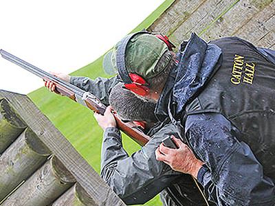 A man aiming a shotgun into the sky with an instructor looking over his shoulder