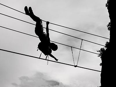 A person hanging upside down from a set of cables, silhouetted against a grey sky