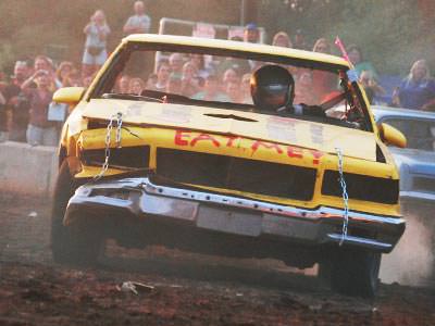 A yellow demolition derby stock car being driven towards the camera, with a crowd in the background