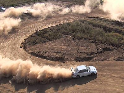 A white car driving around a hairpin bend on a dirt road, kicking up dust behind it