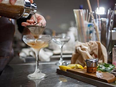 A man's hand pouring a cocktail through a shaker and into a martini glass, with utensils and ingredients on the side