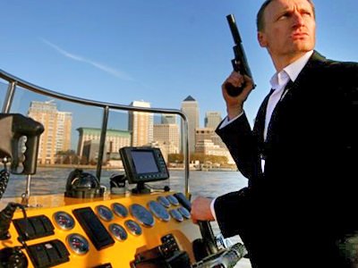 A man in a black suit, holding a handgun whilst driving a boat 
