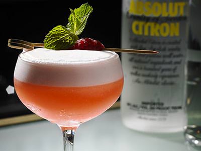 An orange, foamy cocktail garnished with a raspberry and mint sprig with a bottle of vodka in the background
