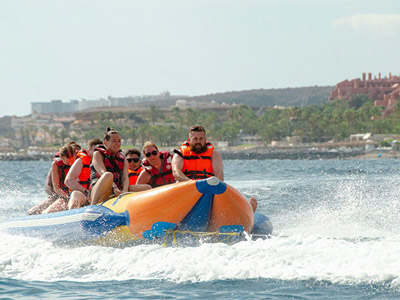 A line of people sat on a banana boat, being pulled by a white boat in the sea