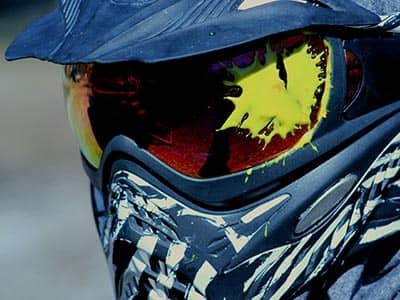 A man's paintball mask with paint splattered on the lens