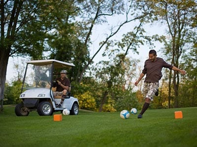 A man kicking a football on a golf course, with a man looking on in a golf buggy in the background