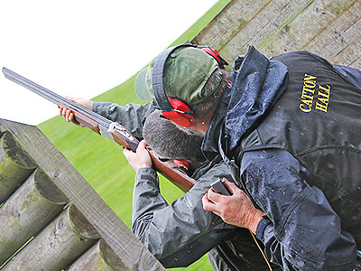 The back of a man aiming with a shotgun with an instructor looking on behind him