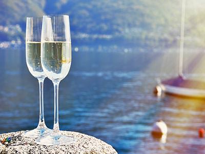Close up of two flute glasses filled with champagne on a rock with a small yacht and boys on water in the background