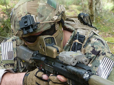 Someone dressed in camouflage outside, aiming a paintball gun