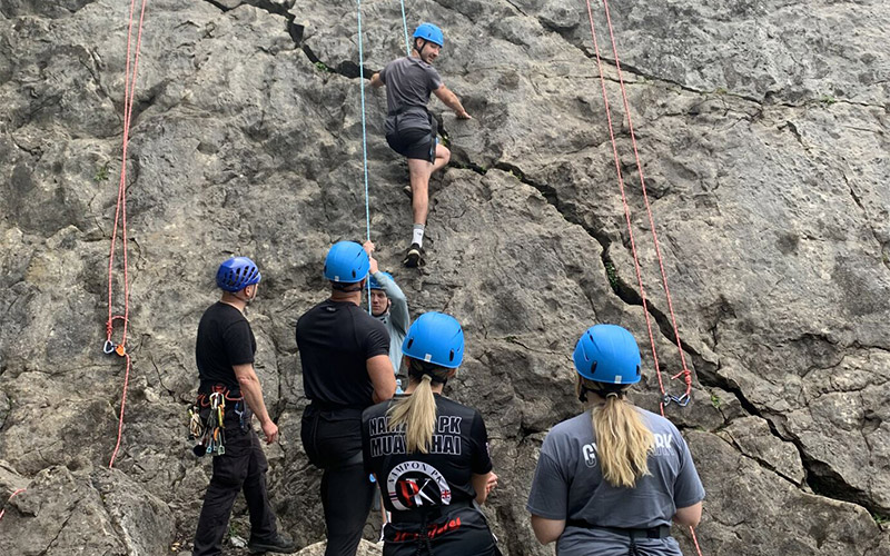 A man rock climbing as a group of people in harnesses watch on 