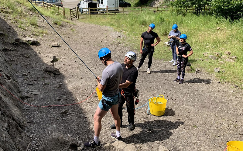 A rock climbing instructor putting a man into a harness whilst three people watch on 