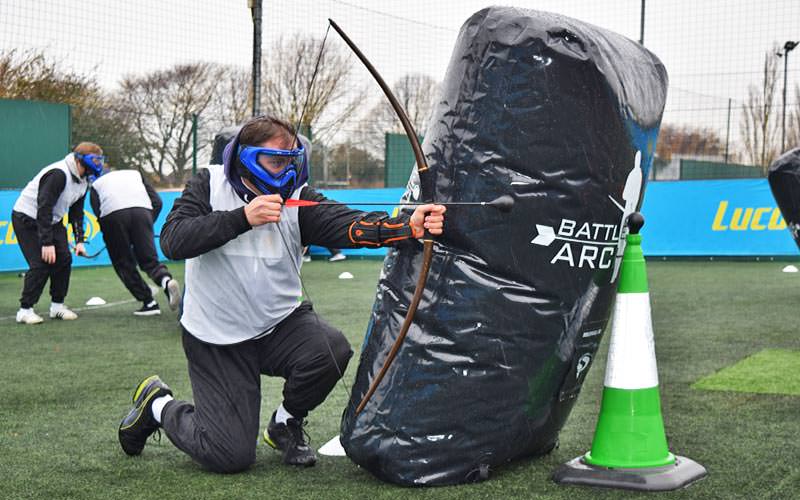 A man wearing a mask and overalls aiming a battlezone archery bow and arrow from behind an inflatable