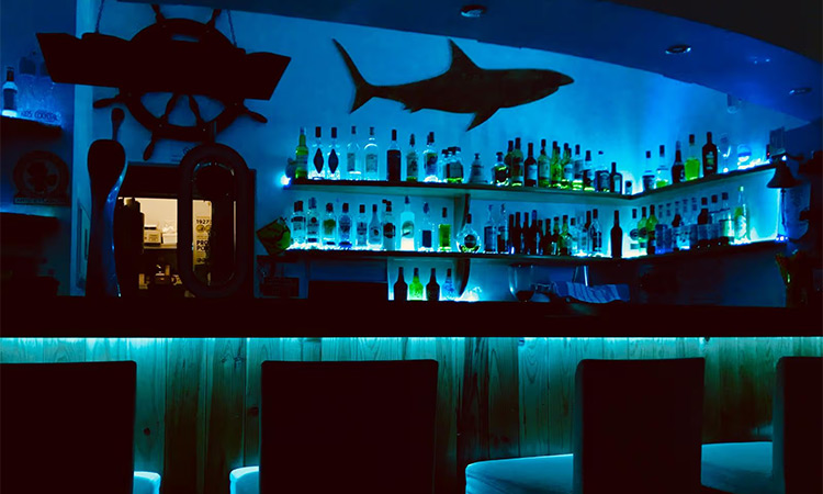 The bar at Sharky's Bar which is in a deep blue light with a shark artwork on the wall as well as a steering wheel from a ship 