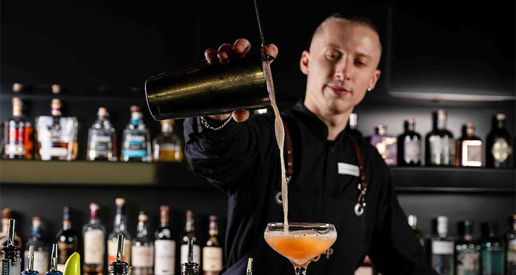 A male bartender in a black work uniform stood at a bar pouring an orange cocktail from a cocktail shaker 