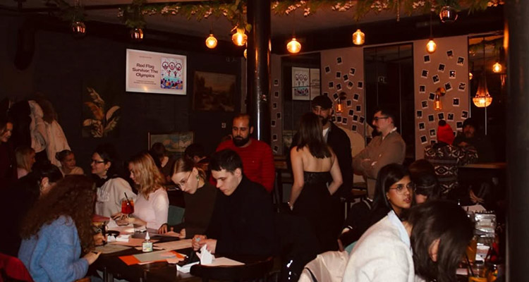 Tables of people drinking at Sloshed Bar which has dim lighting and Polaroid pictures on the wall