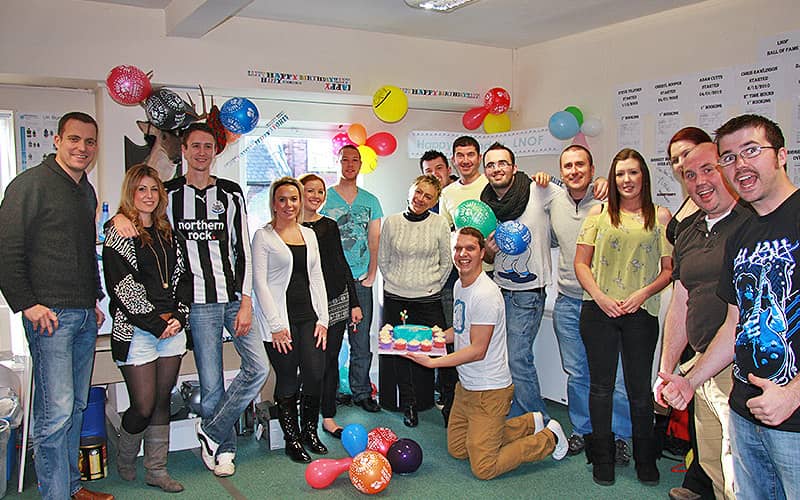 A group of men and women posing with balloons, with one man on his knees on the floor holding a cake 