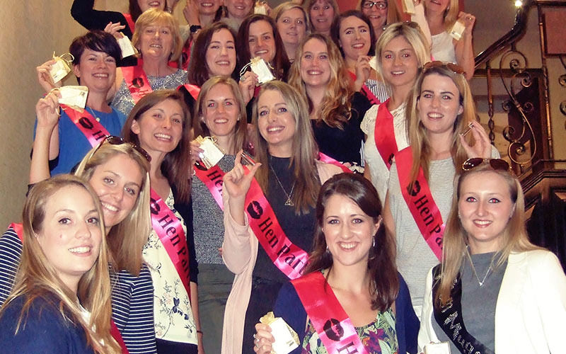 A group of girls all wearing hen party sashes and standing on stairs