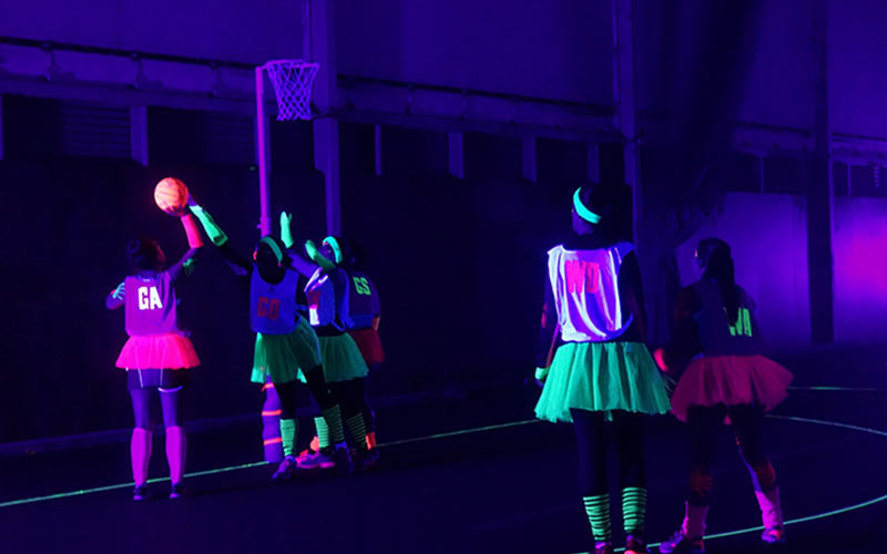 A group of women playing netball in the dark, wearing glow in the dark clothes