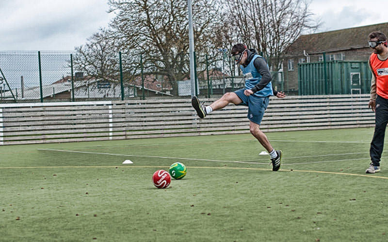 A man trying to kick a ball whilst handicapped by the binocular goggles
