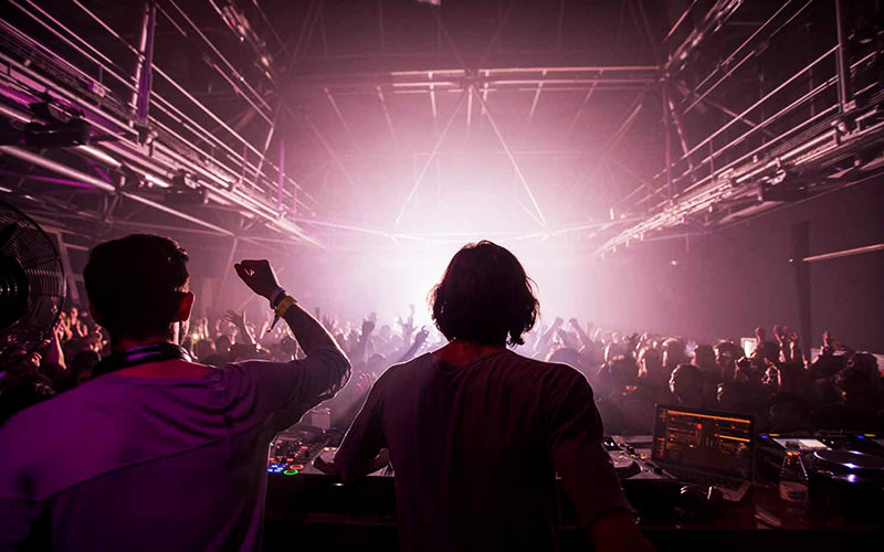 Two DJs performing to a dark room with silouettes of people