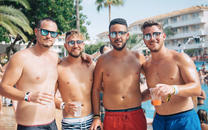Four men wearing swimming shorts by the side of the pool, holding their drinks