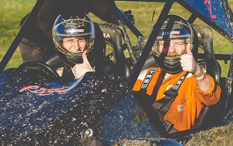 Two men in a muddy kart with their thumbs sticking up