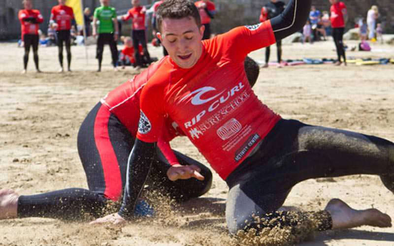 Some men wearing wetsuits and T-shirts, skidding on the sand