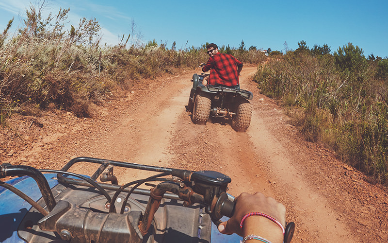 A close up of someone riding on a quad bike, with another man riding in front of him, on a dirt track