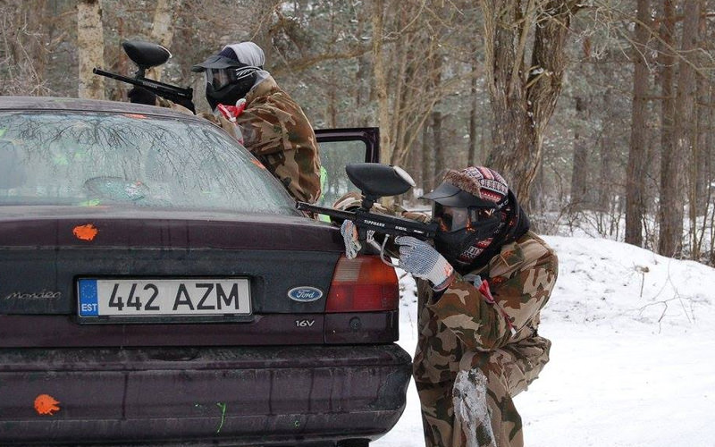 Two people dressed in camouflage hiding behind a car and carrying paintball guns