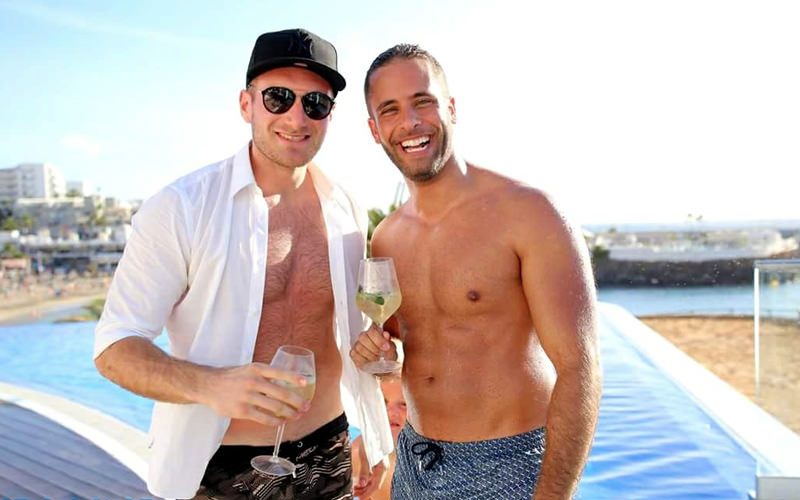 Two men holding drinks with a swimming pool and beach in the background