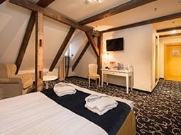 Close up of double bed with blue runner and high wooden beamed ceiling with a chair and tv mounted on wall