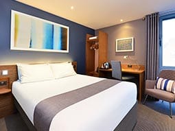 Close up image of a room with a double bed with grey curtains and a grey bed runner with wooden furniture and a grey chair with a striped cushion 