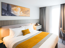 Image of a room with a double bed with a grey headboard and a yellow bed runner and a grey fabric chair and small round glass table next to a window with grey curtains 