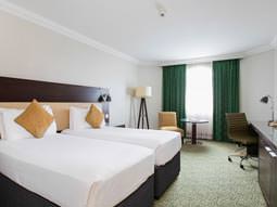 Image of a room with two single beds with white bedding with green curtains and a light green carpet, a floor lamp in the corner of the room with a chair and small round table