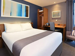 A spacious Travelodge super room with a double bed, desk space. a chair and lots of storage at Travelodge London Docklands