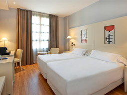 A twin room with two double beds, a desk and chair, bedside lamps and artwork above the beds at NH Valencia Las Artes 