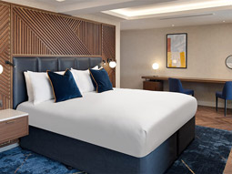 A blue themed double bed against a wooden wall with a long desk and two chairs at the side at Hilton London Metropole