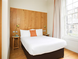 A double bed against a wooden headboard with two bedside lamps and a large window at Cairn Hotel in Edinburgh