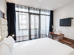 A king bed in a room with a tv on the wall in front of it and a large set of glass sliding doors that lead onto a balcony at Petit Palace Cliper Gran Via Hotel in Madrid 