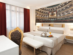 A king bed with an canvas of a theatre behind the bed and a desk and chair in front at Hotel Mayorazgo in Madrid 