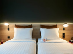 Two single beds against a headboard with warm lighting and a dark wall at Moxy Chester