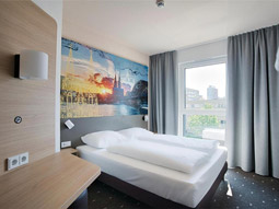 A double bed in a room with large windows and a desk area at B&B Hotel Cologne City