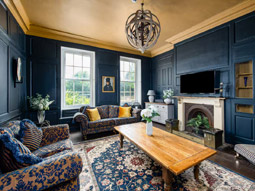 The living room of Leonora House with has deep blue panelling, patterned sofas, a period fireplace and a wall-mounted flatscreen TV 
