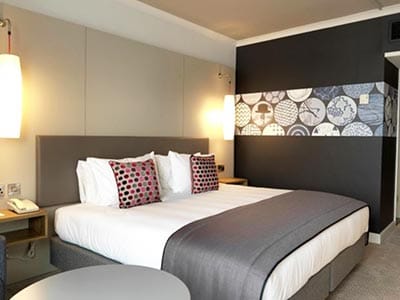 Image of a double bed with grey bed runner and a grey head board and a black wall with grey circle print and two lights attached to the wall each side of the bed