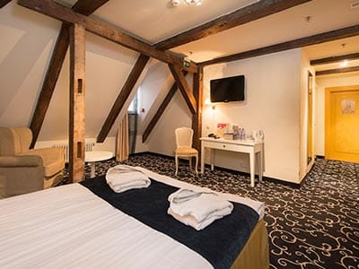 Close up of double bed with blue runner and high wooden beamed ceiling with a chair and tv mounted on wall