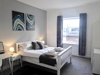 Image of a rooom with a white wood double bed and a window with grey curtains with white furniture