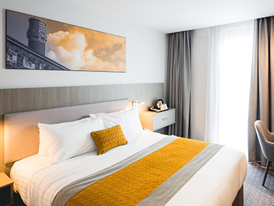 Image of a room with a double bed with a grey headboard and a yellow bed runner and a grey fabric chair and small round glass table next to a window with grey curtains - Rank #5