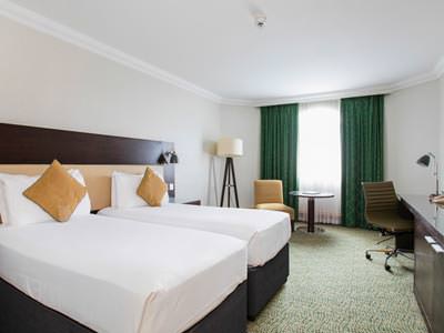 Image of a room with two single beds with white bedding with green curtains and a light green carpet, a floor lamp in the corner of the room with a chair and small round table 